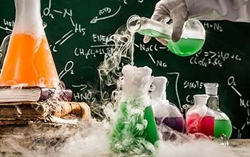 Hand pouring liquid into beaker on a desk. Another beaker on a stack of books with smoke surrounding all beakers and table
