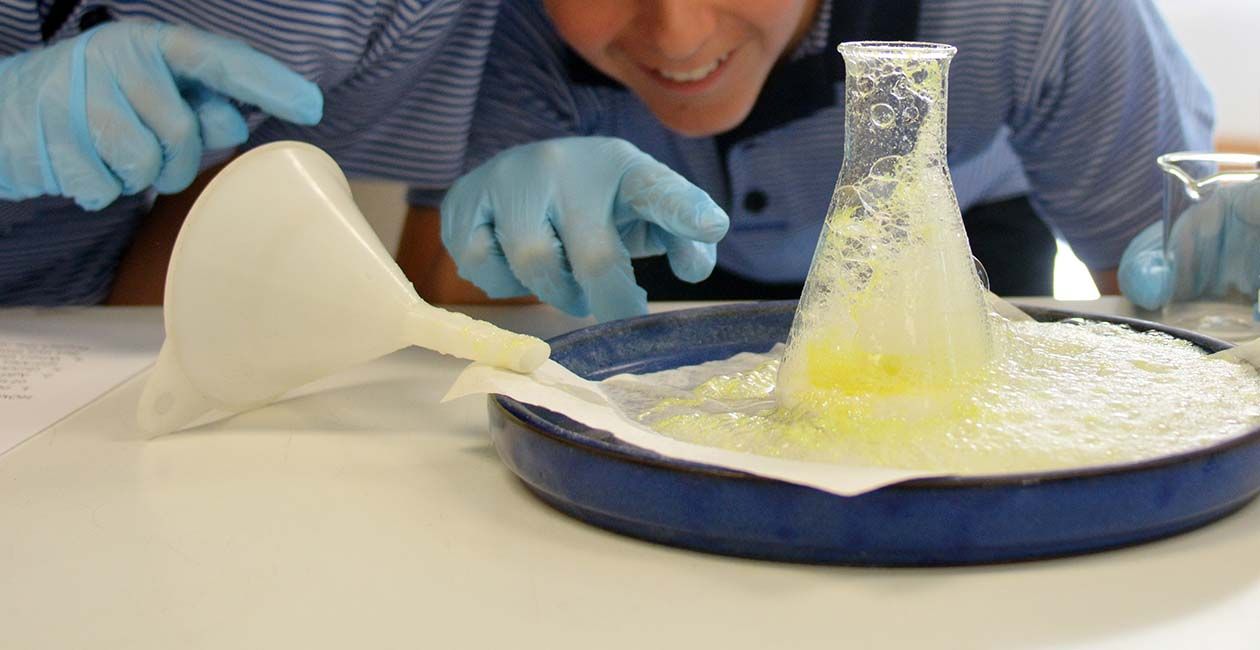 A boy's hand pointing at a beaker with yellow liquid overflowing onto a blue tray